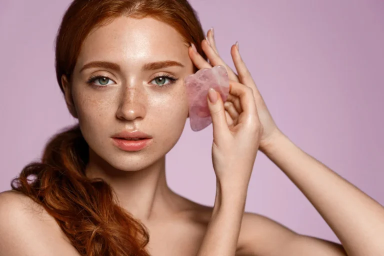 Ragazza con i capelli rossi e le lentiggini su sfondo rosa che usa un Gua Sha